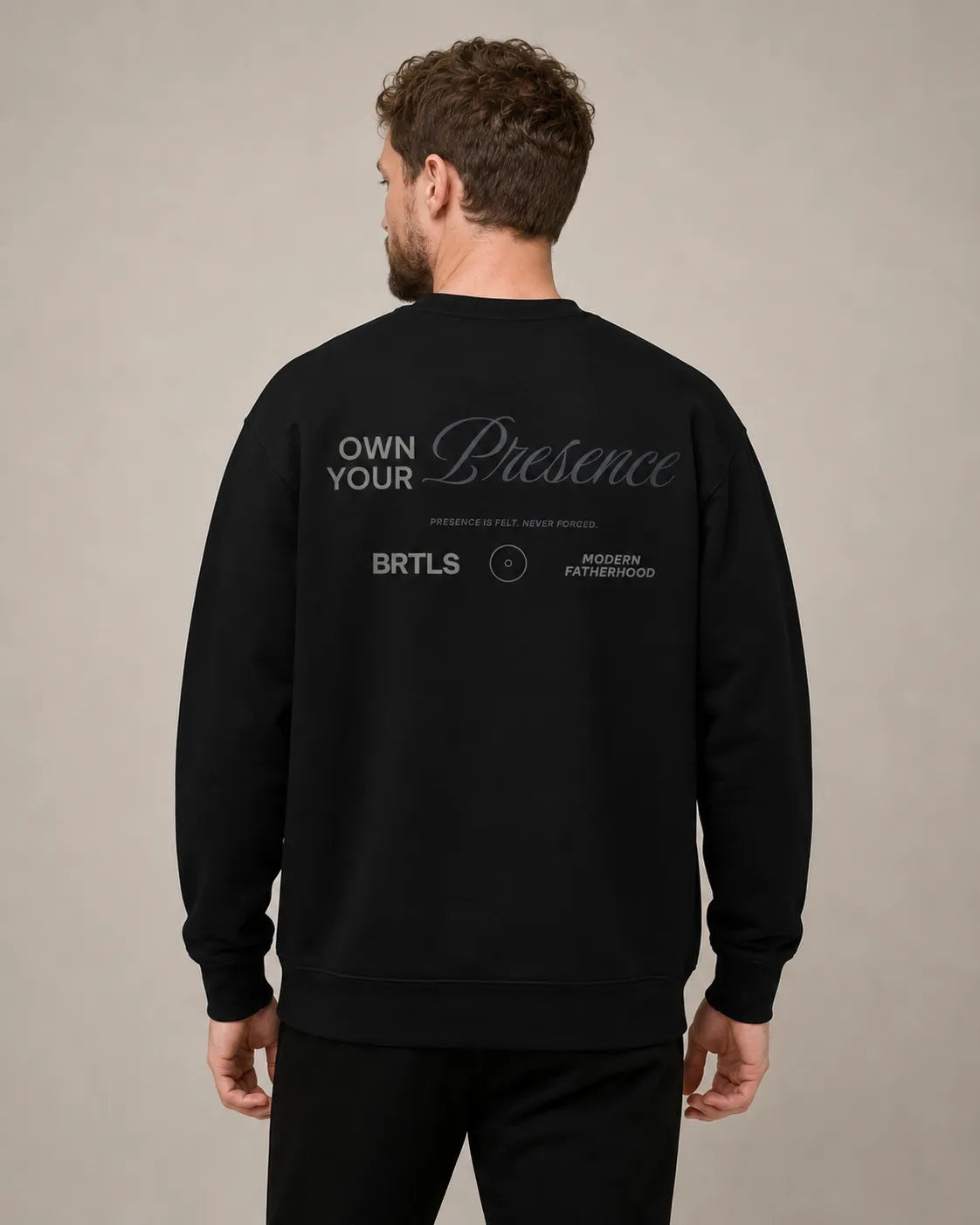 Male model wearing the black BRTLS Own Your Presence sweatshirt showing tonal back artwork in soft editorial lighting