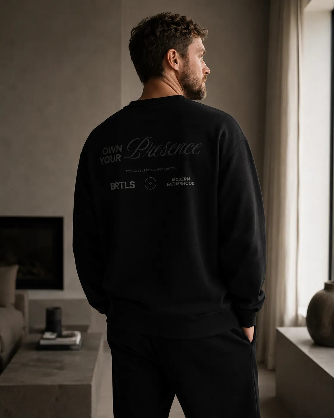 Modern man wearing the black BRTLS Own Your Presence sweatshirt in a refined Scandinavian interior with tonal monochrome styling