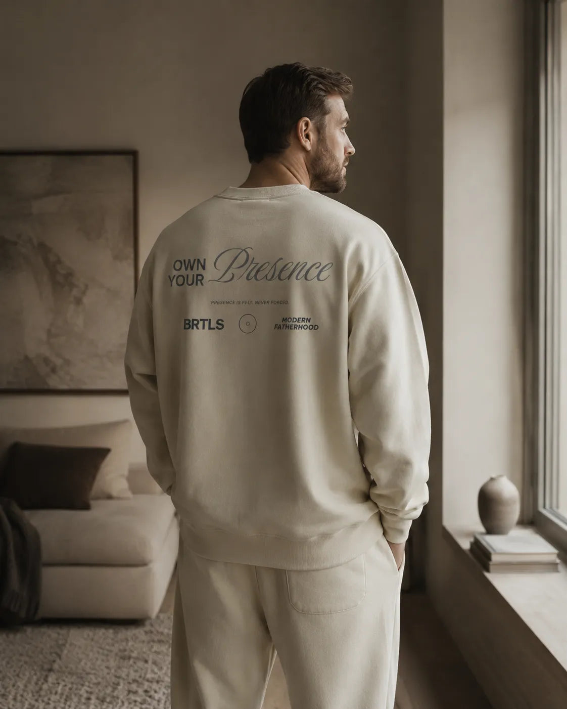 Man wearing the BRTLS Own Your Presence sweatshirt in bone inside a minimalist Scandinavian interior