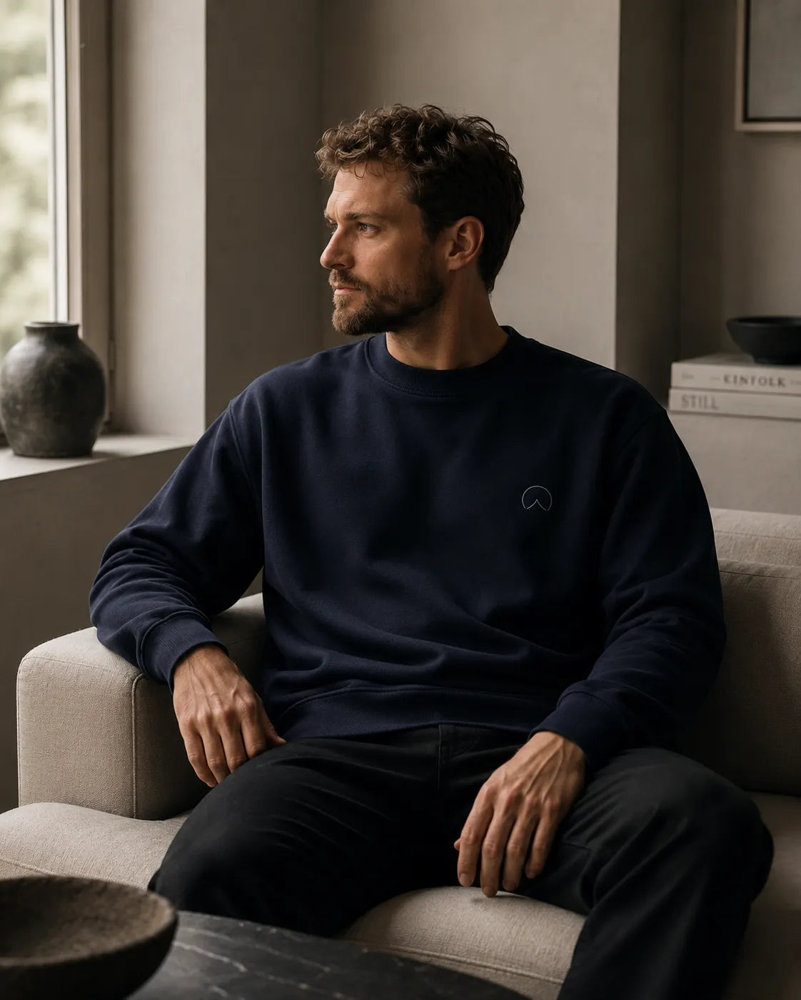 Man wearing the BRTLS Own Your Presence Navy Sweatshirt while seated in a minimalist Scandinavian interior with soft natural light and quiet luxury aesthetic.