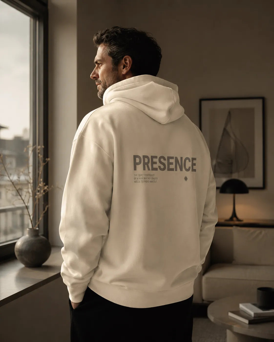 Man wearing the bone BRTLS Presence Hoodie in a warm Scandinavian interior with quiet luxury styling