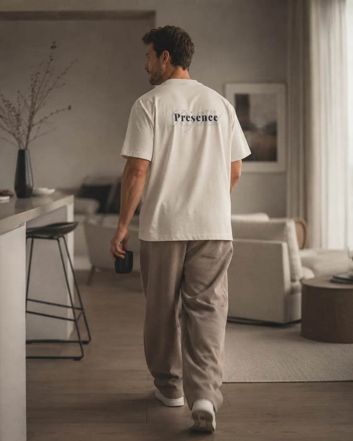 Full body lifestyle image of a man wearing the BRTLS Presence Refined Tee in Vintage White styled within a calm minimalist interior inspired by intentional modern living.