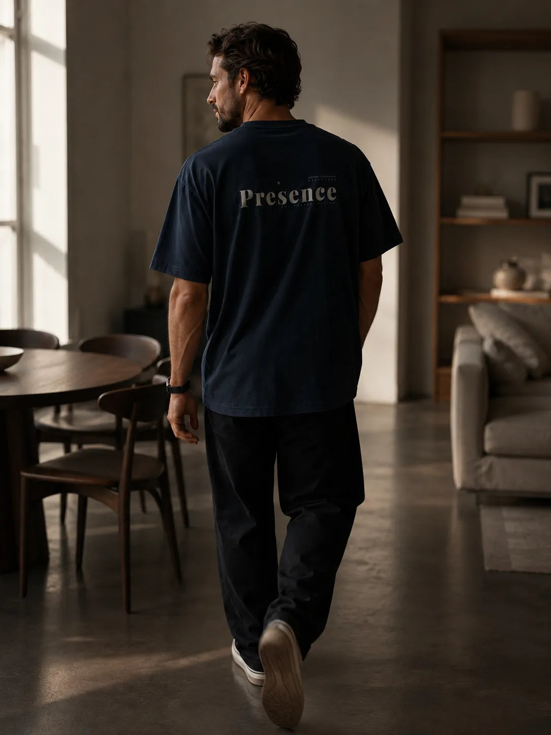 Modern lifestyle image of a man walking indoors wearing the oversized BRTLS Refined Presence Tee in Navy Blazer with tonal artwork